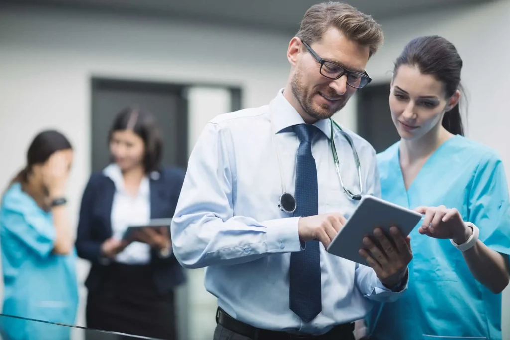 A-male-doctor-in-a-white-shirt-and-tie-is-discussing-information-on-a-digital-tablet-with-a-female-nurse-in-blue-scrubs. In-the-background,-two-other-medical-professionals-are-engaged-in-conversation. The-scene-represents-the-use-of-digital-tools-in-healthcare-for-improved-communication-and-SEO-in-Healthcare.