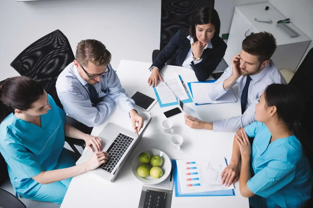 SEO-in-Healthcare - Five healthcare professionals are gathered around a white table in a modern meeting room. Two are in medical scrubs, while the other three are in business attire. They are discussing documents, charts, and a laptop screen. Green apples are placed in a bowl on the table. One person is pointing at the laptop, another is on the phone, and the rest are actively engaged in the discussion.