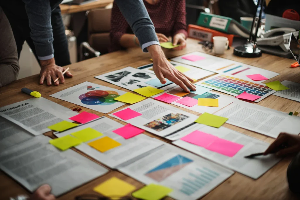 A group of individuals are collaborating on a wooden table covered with various digital marketing strategy materials. The table displays an array of colorful sticky notes in yellow, green, pink, and blue, alongside printed documents with text and images. There are also color swatches and a digital tablet displaying graphs. The hands of the individuals are seen pointing and placing sticky notes, indicating an active planning session focused on developing digital marketing strategies.