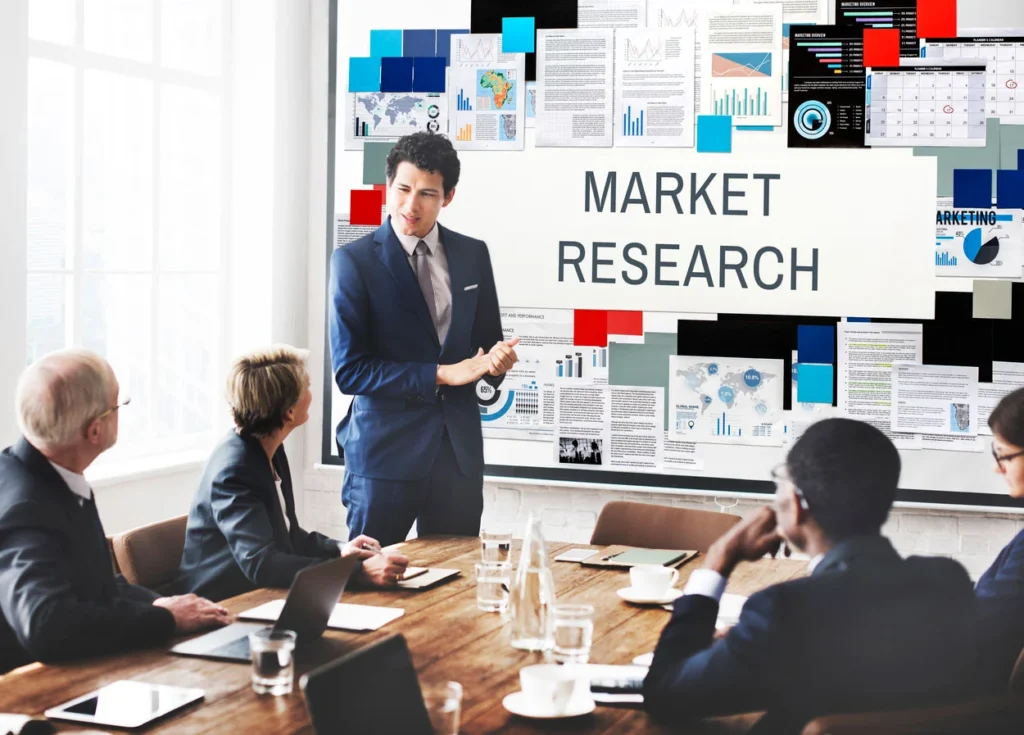 A business professional in a blue suit presents digital marketing strategies and market research data to a diverse group of colleagues in a meeting room. The presentation board behind him displays various charts, graphs, and maps, along with the prominent title "MARKET RESEARCH." The colleagues are seated around a conference table with laptops and documents in front of them, attentively listening to the presentation.