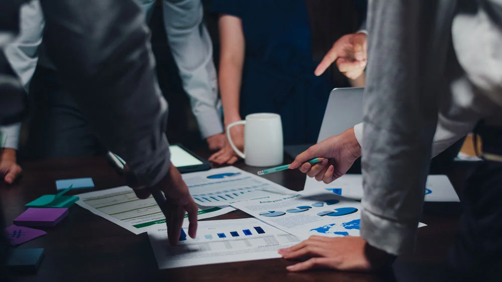 Business professionals collaborate around a table covered with documents and charts, discussing digital marketing strategies and market research data. Their hands and gestures indicate a dynamic conversation, with various data points and graphs visible on the papers.